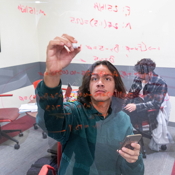 A male student writes equations on a transparent dry erase board. 