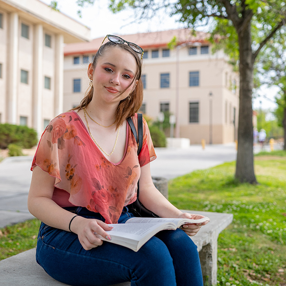 A female student reading a textbook outside on a bench on the NMSU campus