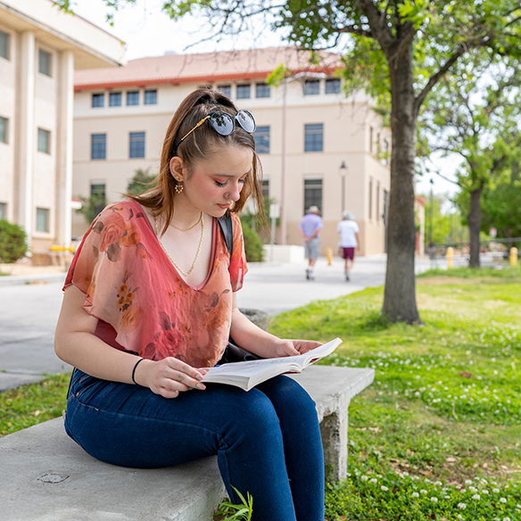 a female student sitting on a bench outside on campus reading a book