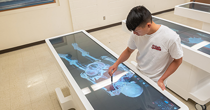 A student uses a large interactive touchscreen table displaying a 3D digital human anatomy model in a classroom setting.