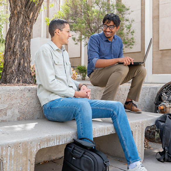Two male students sitting outside on a bench while talking and browsing on a laptop,