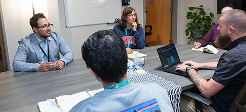 a group of people speaking at a table, referencing a laptop