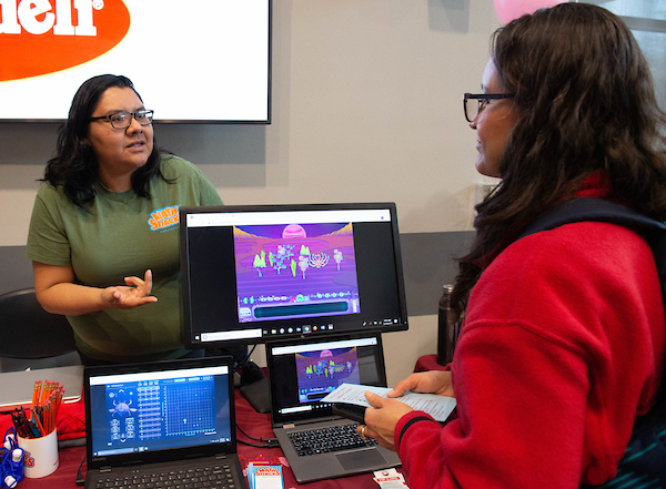 Two women speaking aross from a computer displaying an online game.